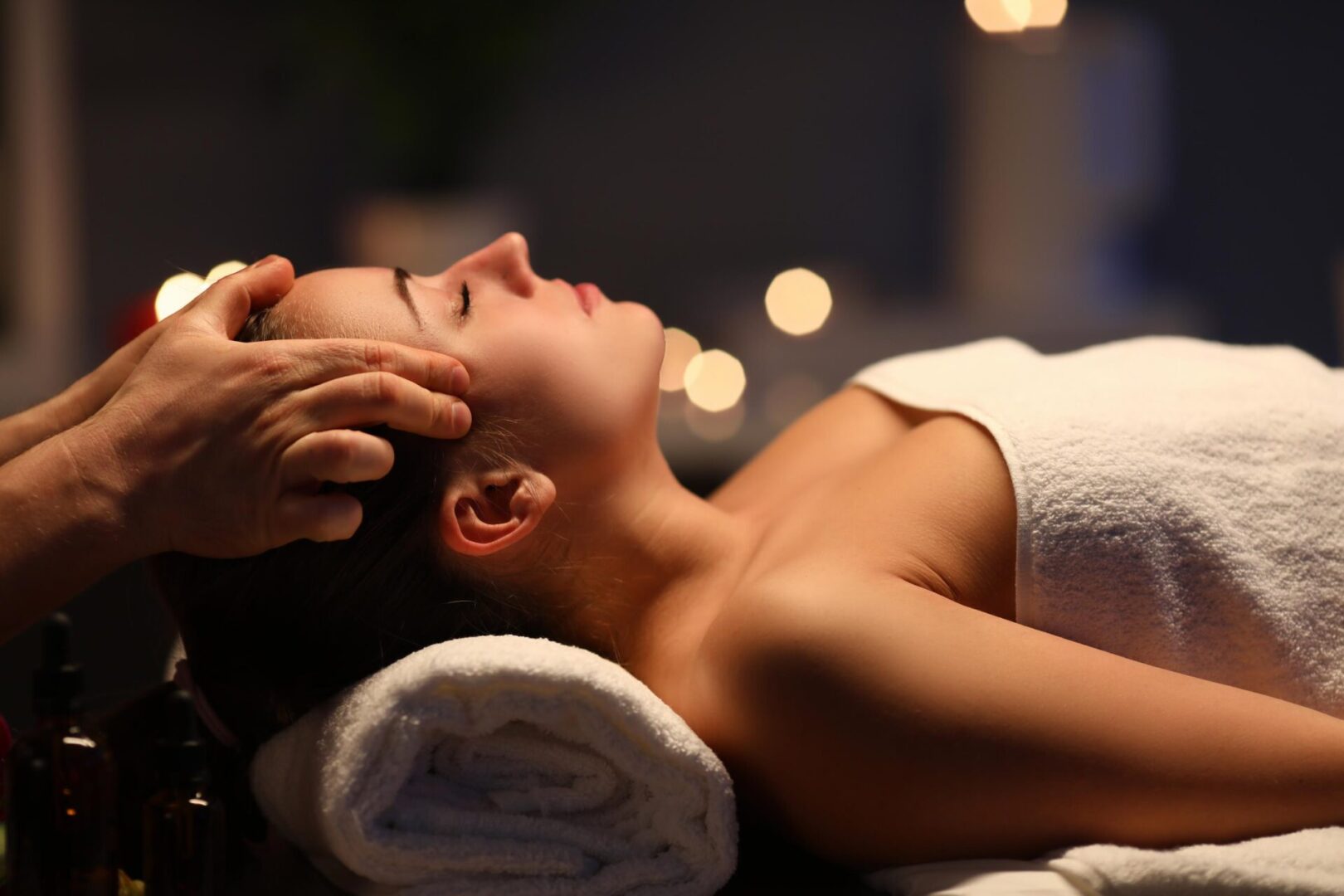 A person receiving a head massage while lying on a towel-covered surface in a dimly lit setting.