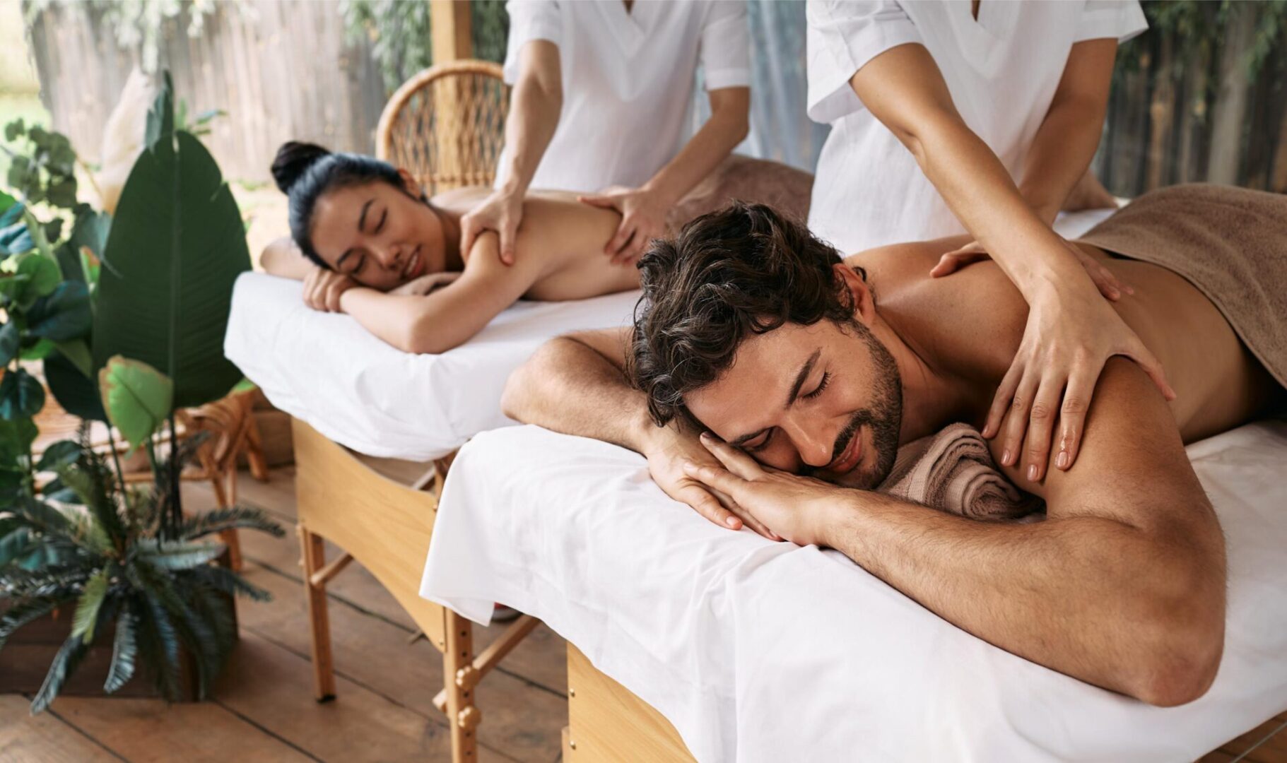 Two people are receiving back massages on adjacent tables in a serene setting with plants, while two massage therapists work.