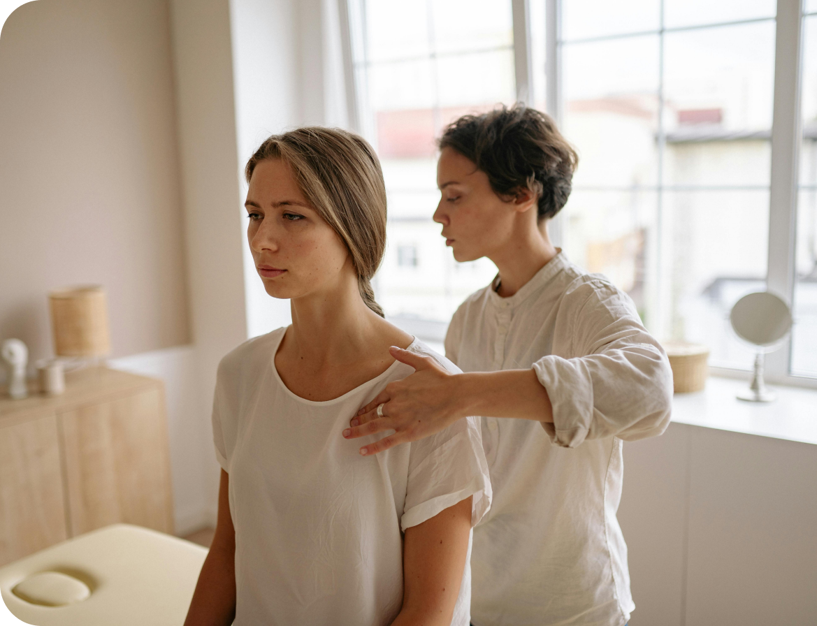 A therapist performs a shoulder examination on a seated woman in a bright room.